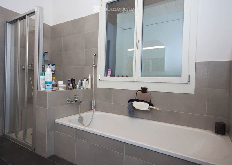 Modern bathroom with a bathtub, shower, and a window. The space features grey tiles, a towel rack, and various bathroom products on the shelf.