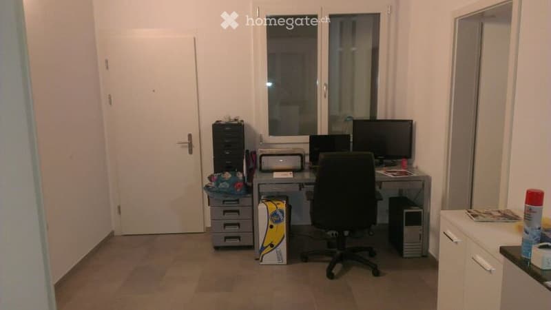 A small, furnished room with a desk, chair, computer, printer, and storage unit. The room has a tiled floor, white walls, and a window with blinds.