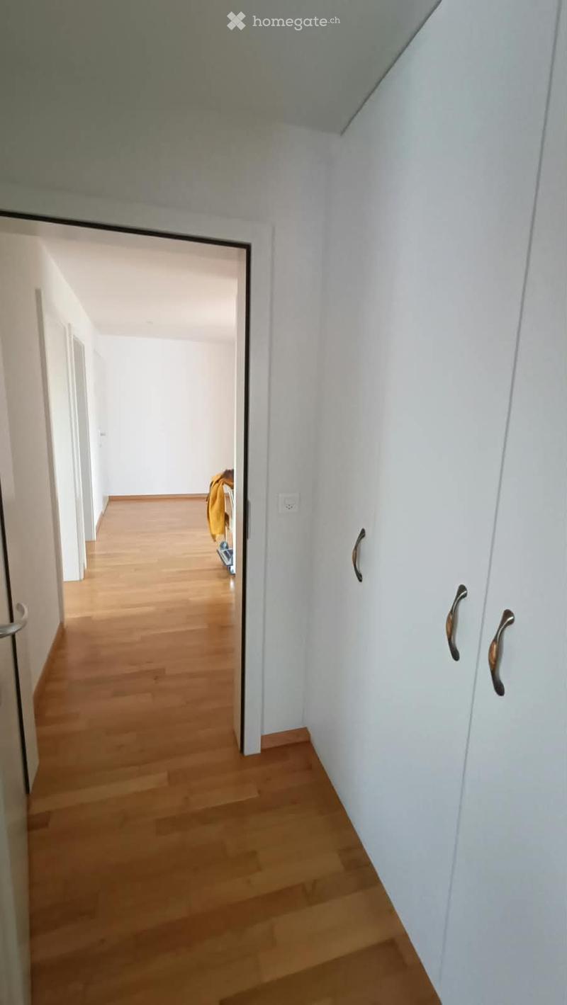 A narrow hallway with light wooden flooring and white walls, featuring a partially open door leading to another room.
