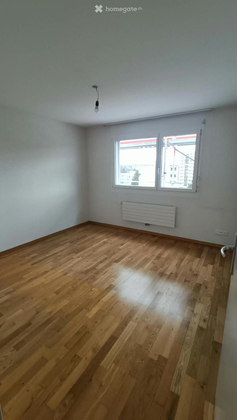 Empty room with wooden flooring, white walls, and a window with a view of buildings outside. A radiator is mounted on the wall below the window.
