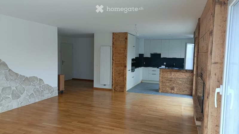 Empty room with wooden flooring, white walls, and a stone accent wall. The room features a doorway leading to a kitchen area with white cabinets and a wooden island.
