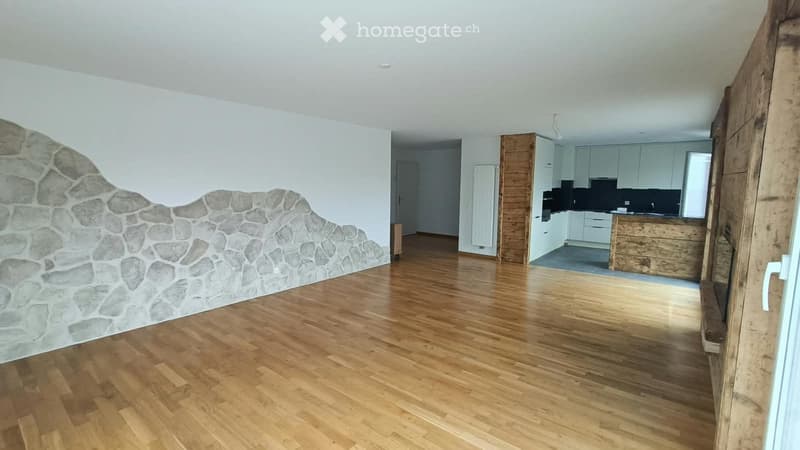 Empty room with wooden flooring and a stone accent wall, leading to a modern kitchen area with white cabinets and stainless steel appliances.