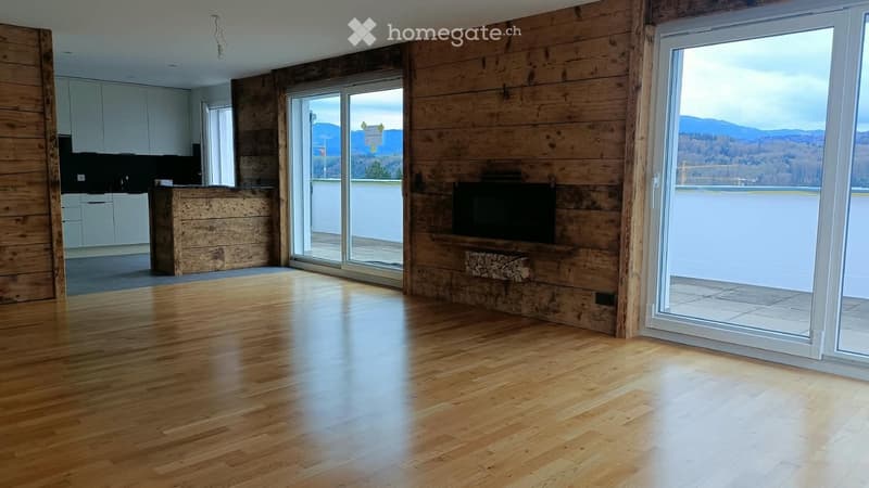 Open-plan living and dining area with wooden flooring, large windows offering a scenic view, a fireplace, and a modern kitchen area with white cabinets.