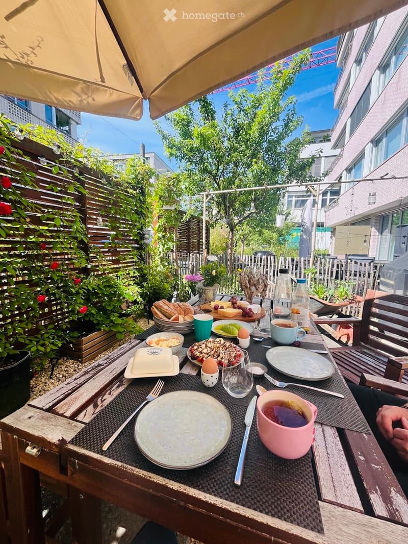 A sunny outdoor dining area with a table set for breakfast, surrounded by greenery and a wooden fence.