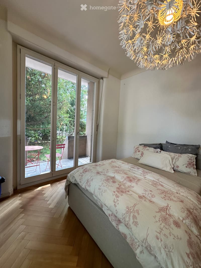 Bright bedroom with wooden flooring, a large bed with floral bedding, a modern chandelier, and a sliding glass door leading to a balcony with outdoor furniture.