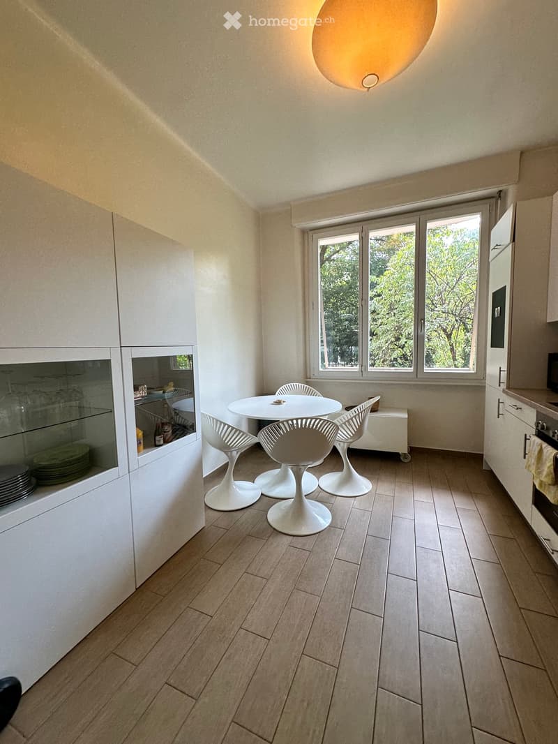 Modern kitchen with white cabinets, a dining area, and a large window overlooking greenery. The space includes a round dining table with four white chairs, a built-in oven, and a dishwasher.