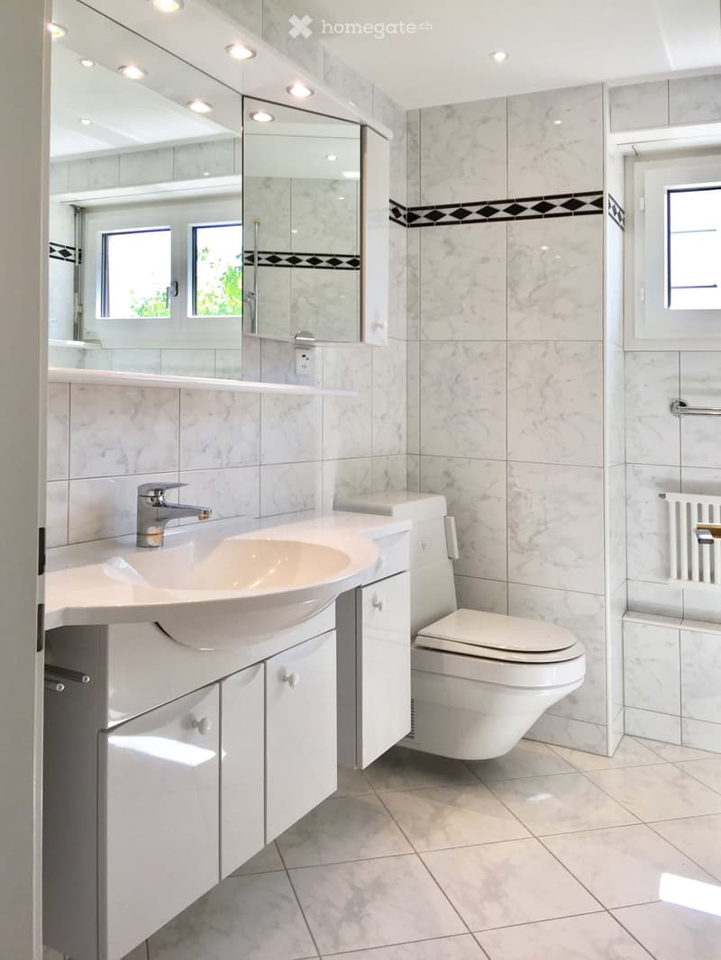 Modern bathroom with white marble tiles, a large mirror, a sink with a modern faucet, and a toilet. The room features recessed lighting and a window with frosted glass.