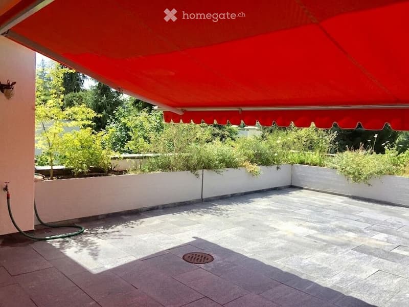 A covered balcony with a red awning, tiled floor, and low white walls with greenery on top.