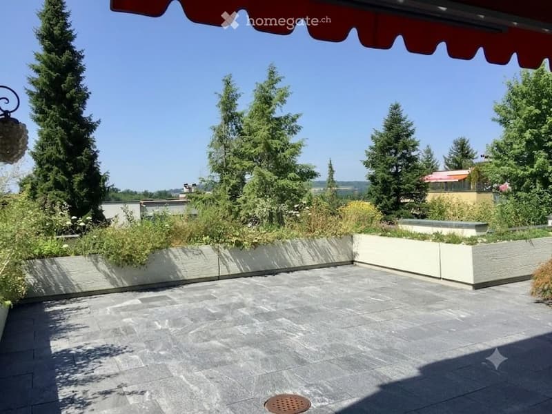 A modern outdoor terrace with a stone-paved floor, raised planters, and surrounding greenery under a red awning.