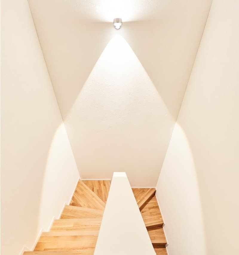 Modern staircase with wooden steps and white walls, illuminated by a single spotlight creating a triangular light pattern on the wall.