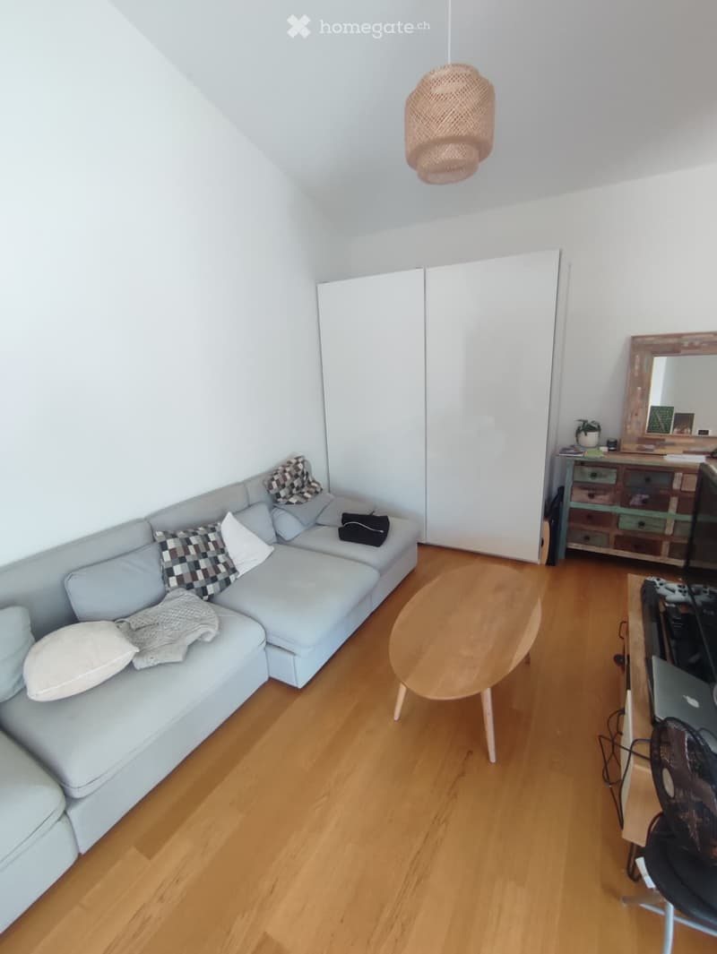 A modern living room with a light blue sectional sofa, wooden coffee table, and a small desk area with a laptop and fan. The room features white walls, wooden flooring, and a large sliding closet.