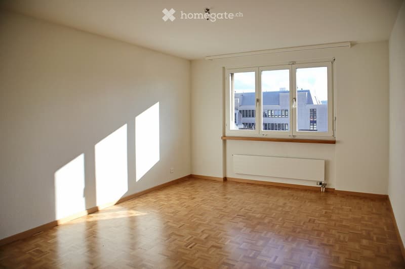 Empty room with wooden flooring, white walls, and large windows allowing natural light to enter. The room features a radiator below one of the windows and a small shelf above it.