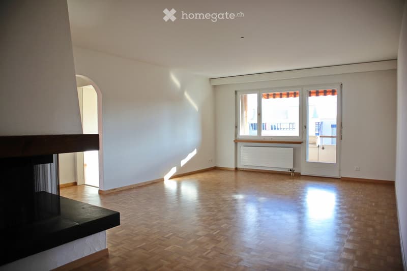 Empty room with wooden flooring, white walls, and a fireplace on the left side. There is a large window with a view and a radiator below it.
