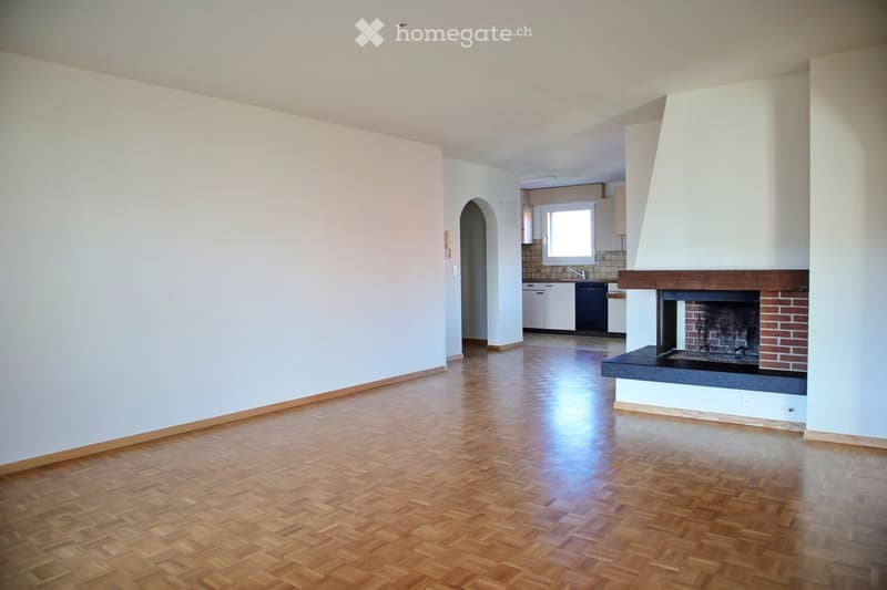 Empty living room with wooden flooring, white walls, and a fireplace. The room is connected to a small kitchen area with white cabinets and a window.