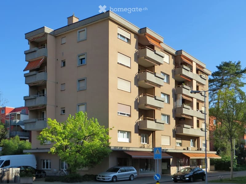 Multi-story residential building with beige facade, multiple balconies, and a parking area in front.