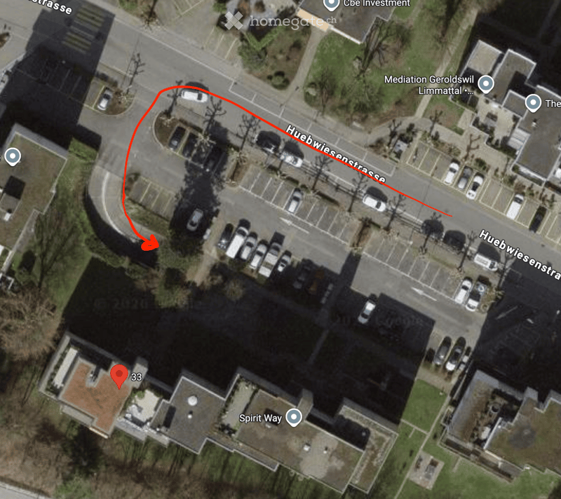 Aerial view of a residential area showing a parking lot, several parked cars, and a building with a red-tiled roof. The area is surrounded by streets and other buildings.