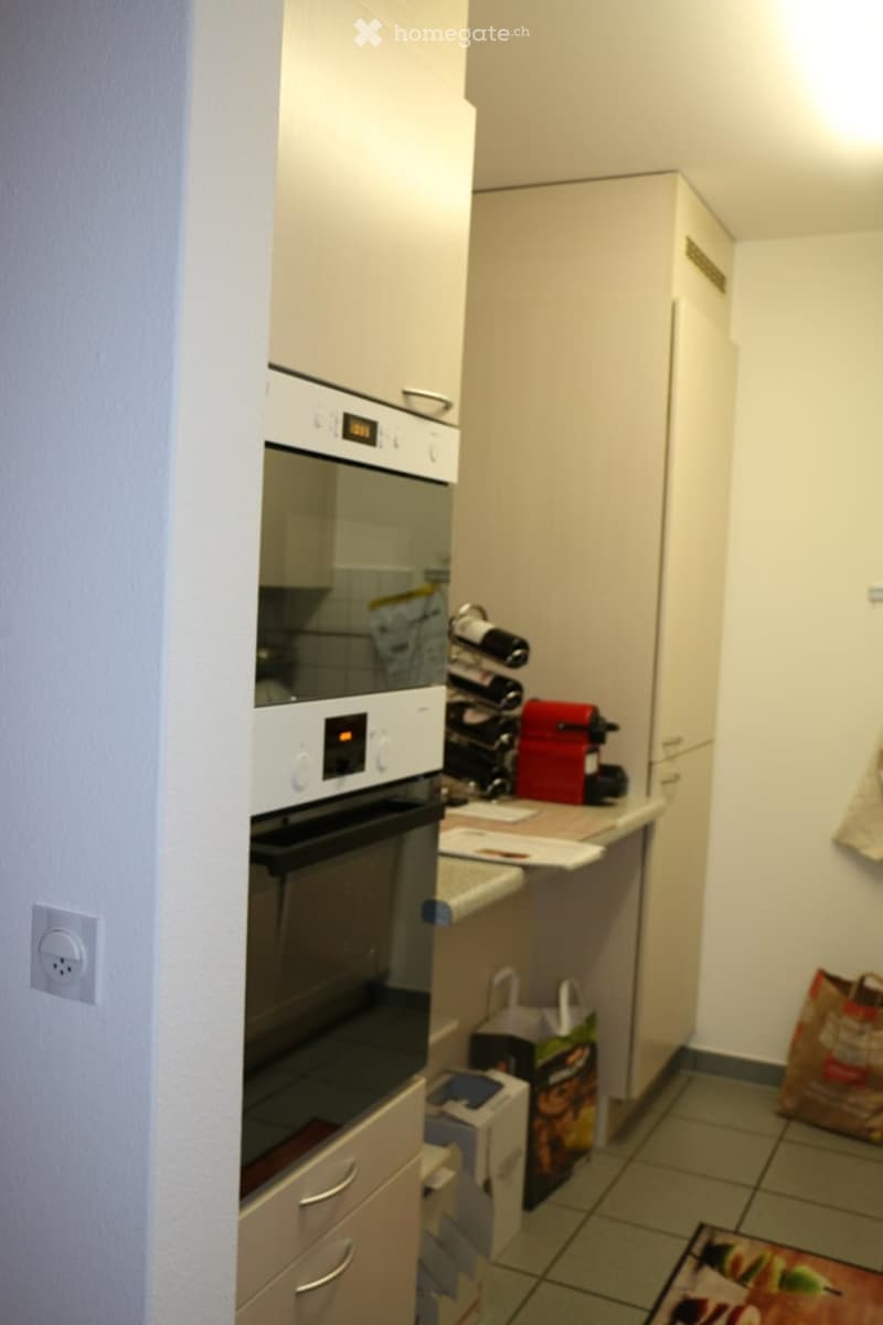 Modern kitchen with white cabinets, built-in oven, wine rack, and countertop. Includes a refrigerator and various items on the counter.