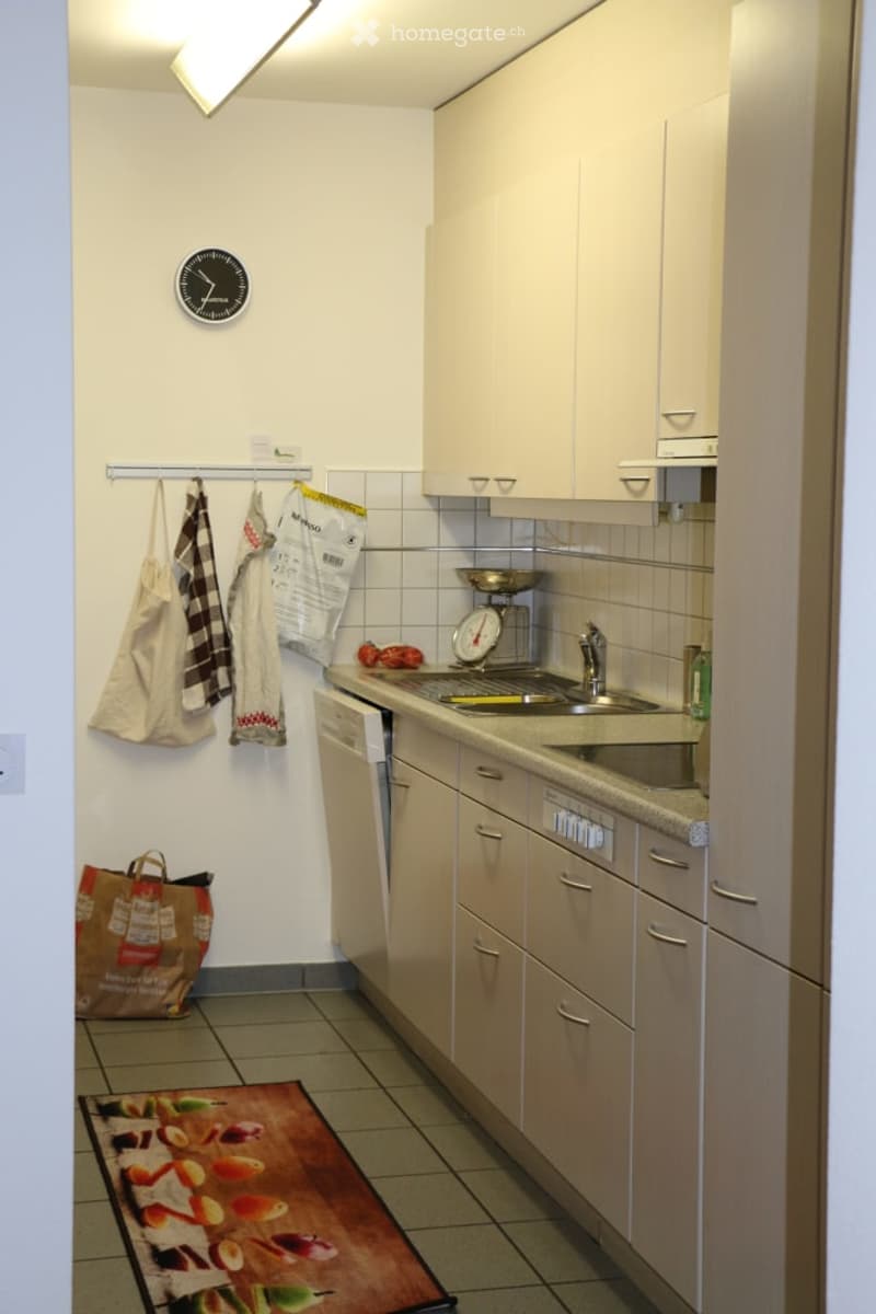 Modern kitchen with light-colored cabinets, a sink, dishwasher, and a scale on the counter. A clock is mounted on the wall, and a shopping bag and handbag are placed on the floor.