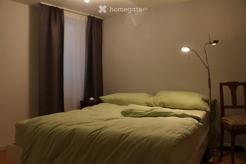 Cozy bedroom with a double bed, green bedding, nightstand, wooden chair, and floor lamp near the window with dark curtains.