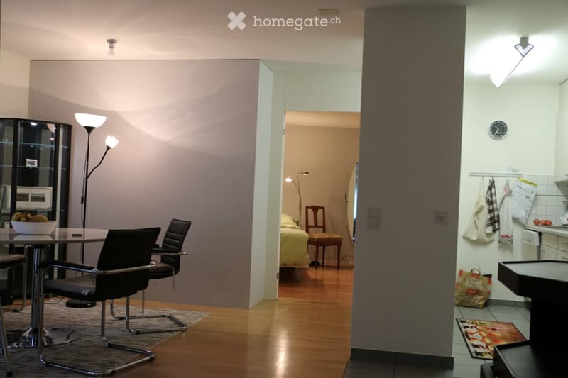 A modern, open-plan living and dining area with a sleek glass table, black chairs, a floor lamp, and a bowl of fruit. A separate bedroom is visible through an open doorway, featuring a bed, a wooden stool, and a mirror. A small kitchen area is also visibl