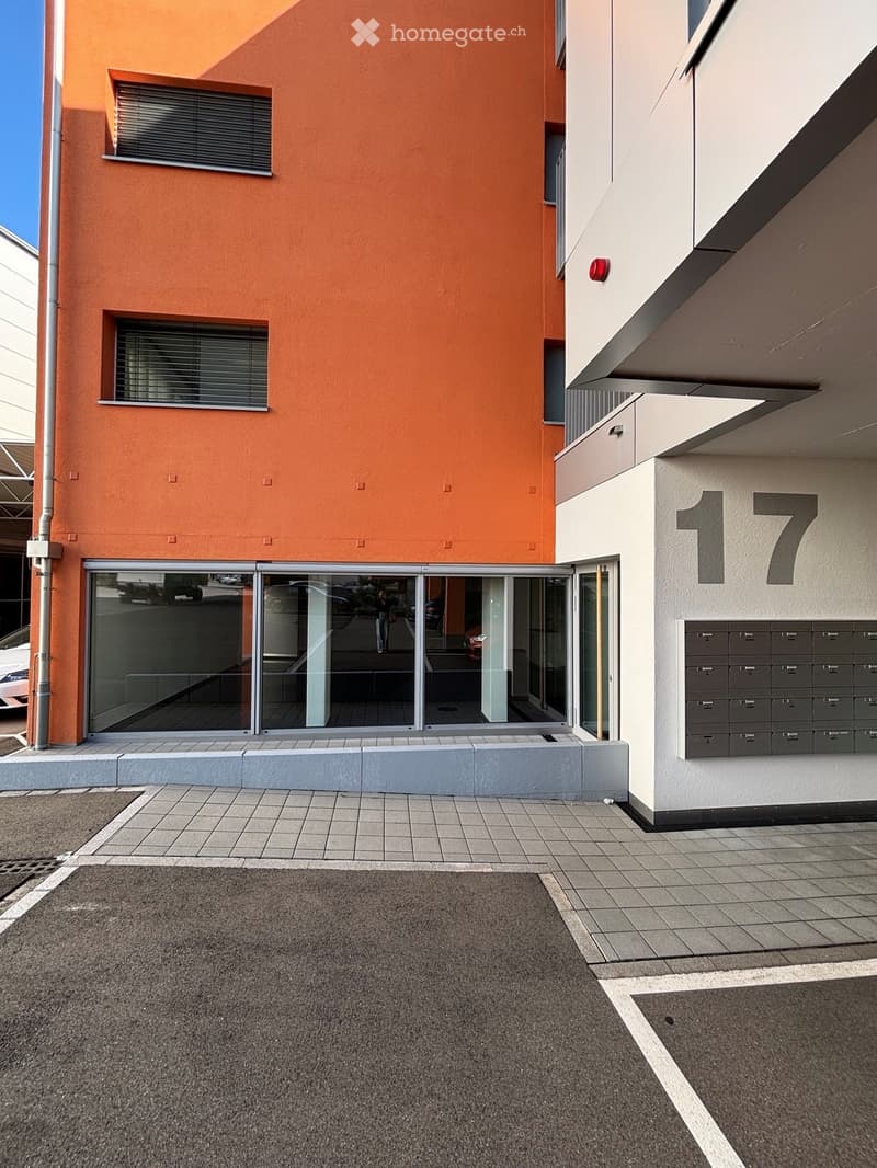 Modern building exterior with orange and white facade, large glass entrance, mailboxes, and tiled pavement.