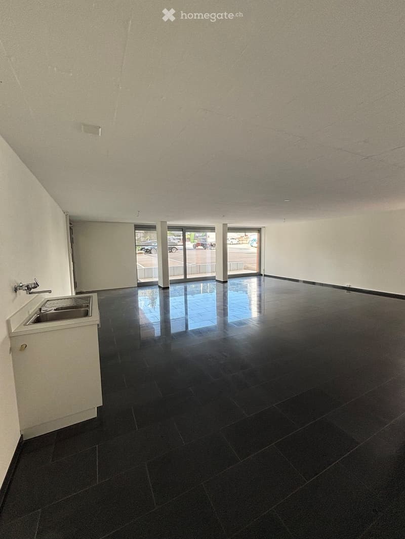 Empty room with black tiled floor, white walls, and a small sink area in the corner. Large glass doors lead to an outdoor area.