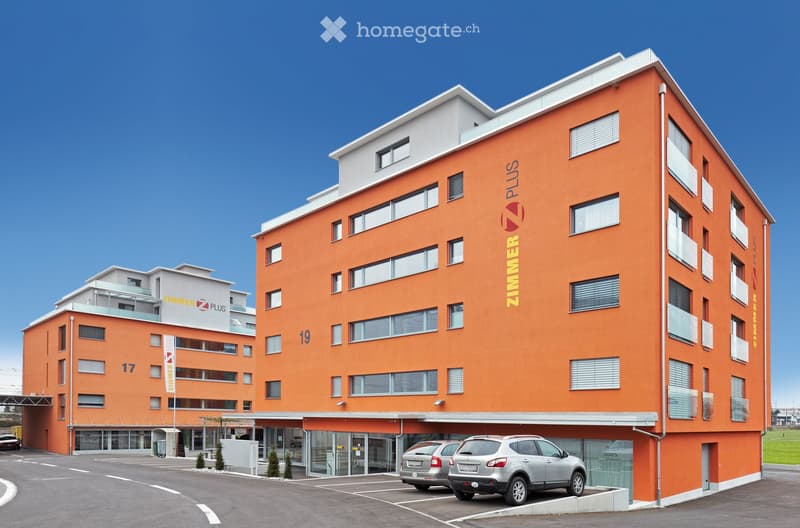 Modern orange apartment building with multiple floors, large windows, and a parking area with several cars.