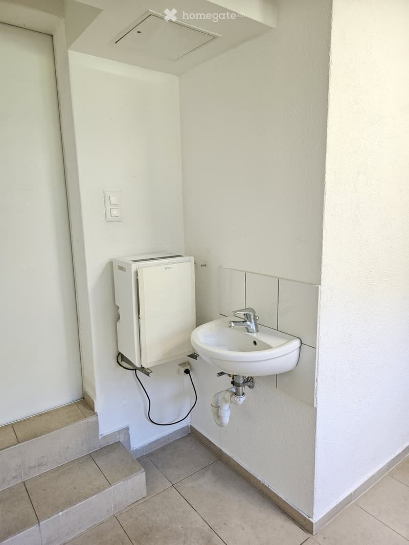 Small bathroom with a white sink, chrome faucet, and a white washing machine mounted on the wall. Light-colored tiled floor and walls.