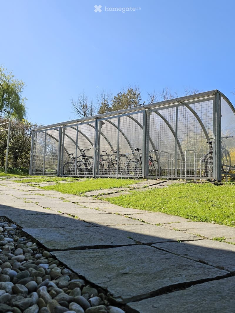 Outdoor area with a covered bicycle shelter containing several bicycles, surrounded by grass and trees.