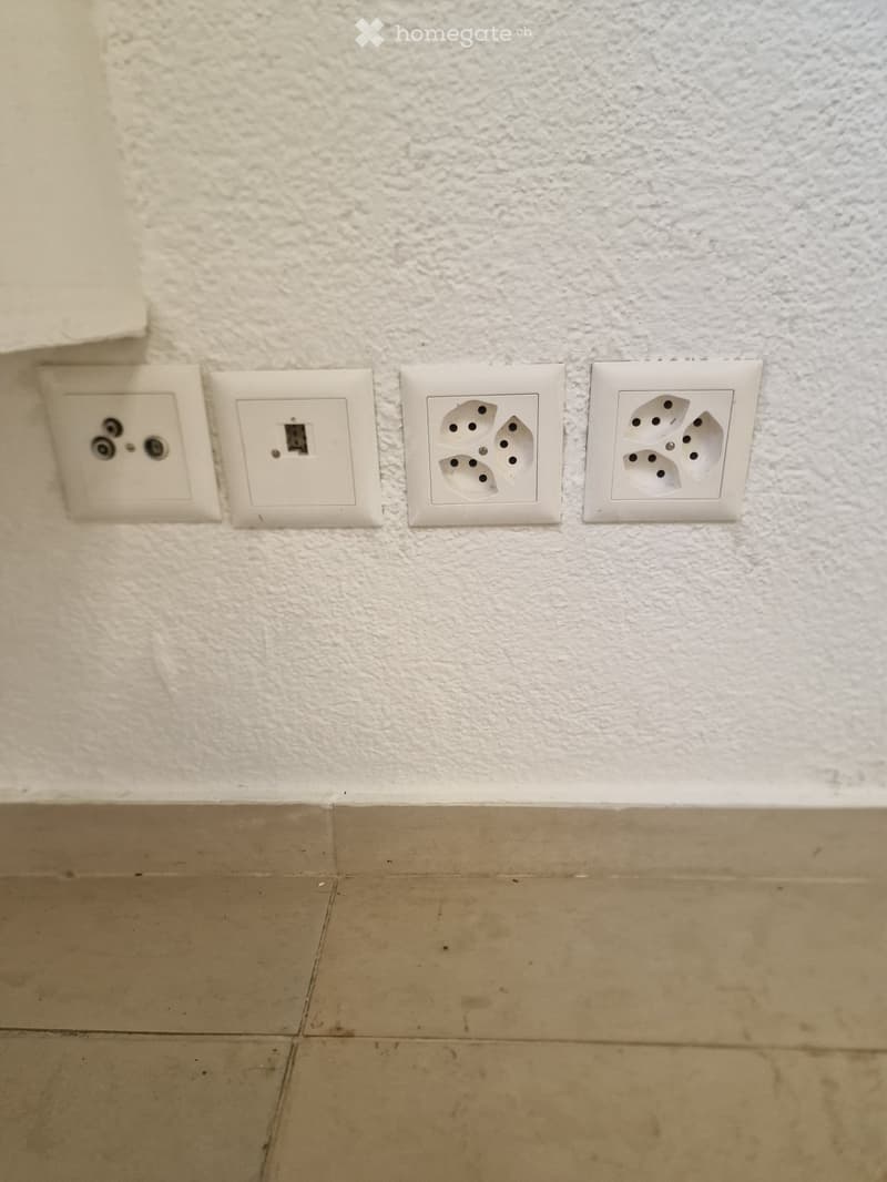 Empty room with beige tiled floor and textured white wall, featuring four electrical outlets and a light switch.