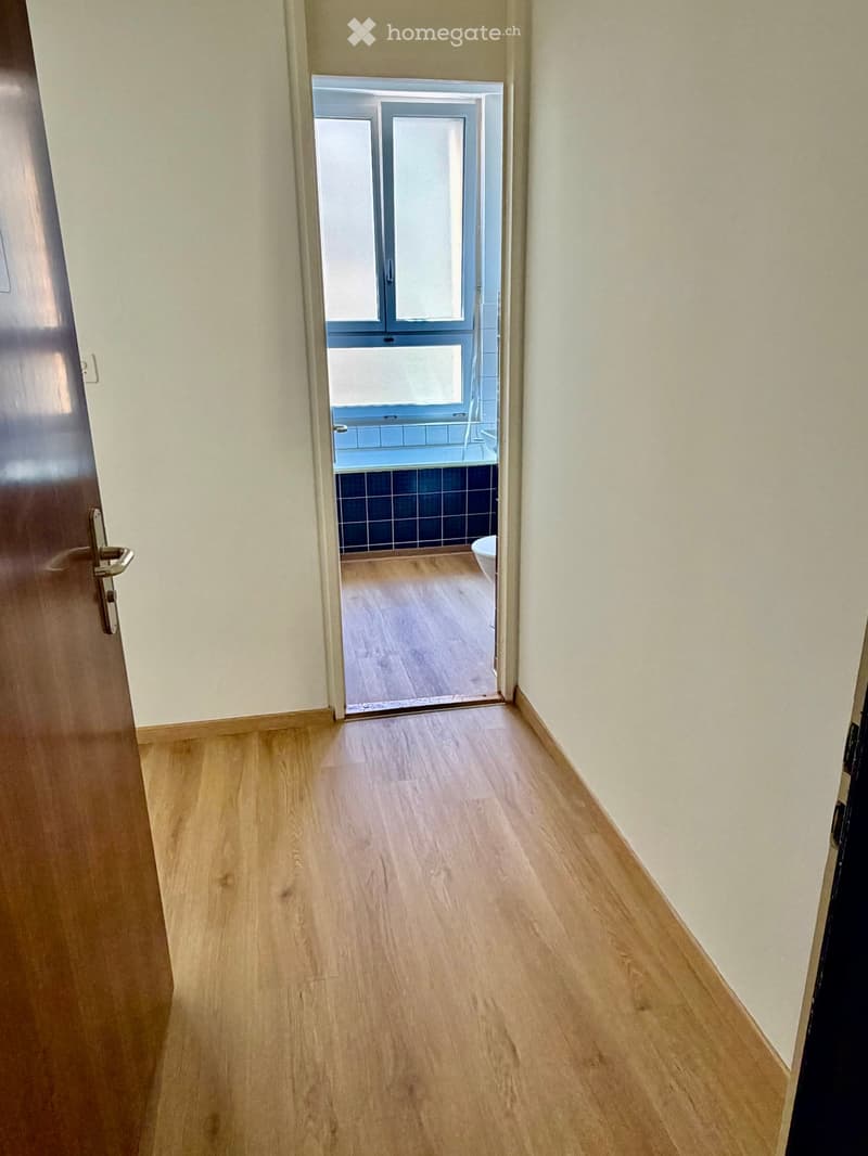 Empty hallway with light wooden flooring and cream-colored walls, leading to a bathroom with blue tiles and a window.