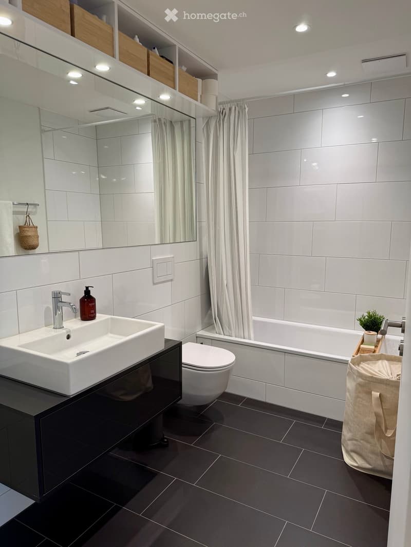 Modern bathroom with white tiles, black floor, white sink, toilet, bathtub, and a large mirror with wooden shelves above.