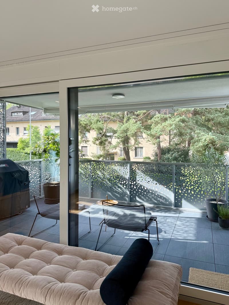 Modern balcony with large glass doors, sofa, chairs, table, potted plants, and a grill cover.