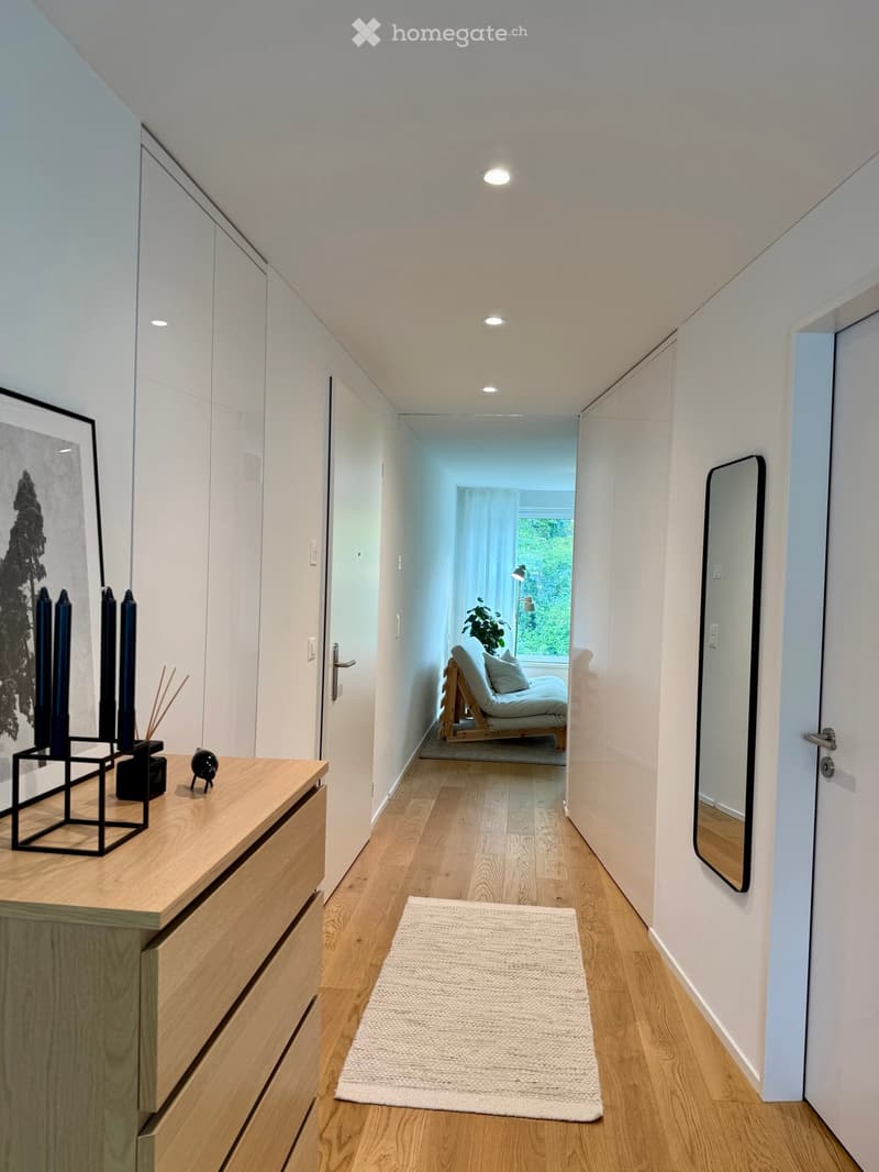 Modern hallway with light wooden flooring, white walls, built-in wardrobes, and recessed lighting. Features a small white rug, a wooden dresser with decorative items, and a view into a room with a daybed and large window.