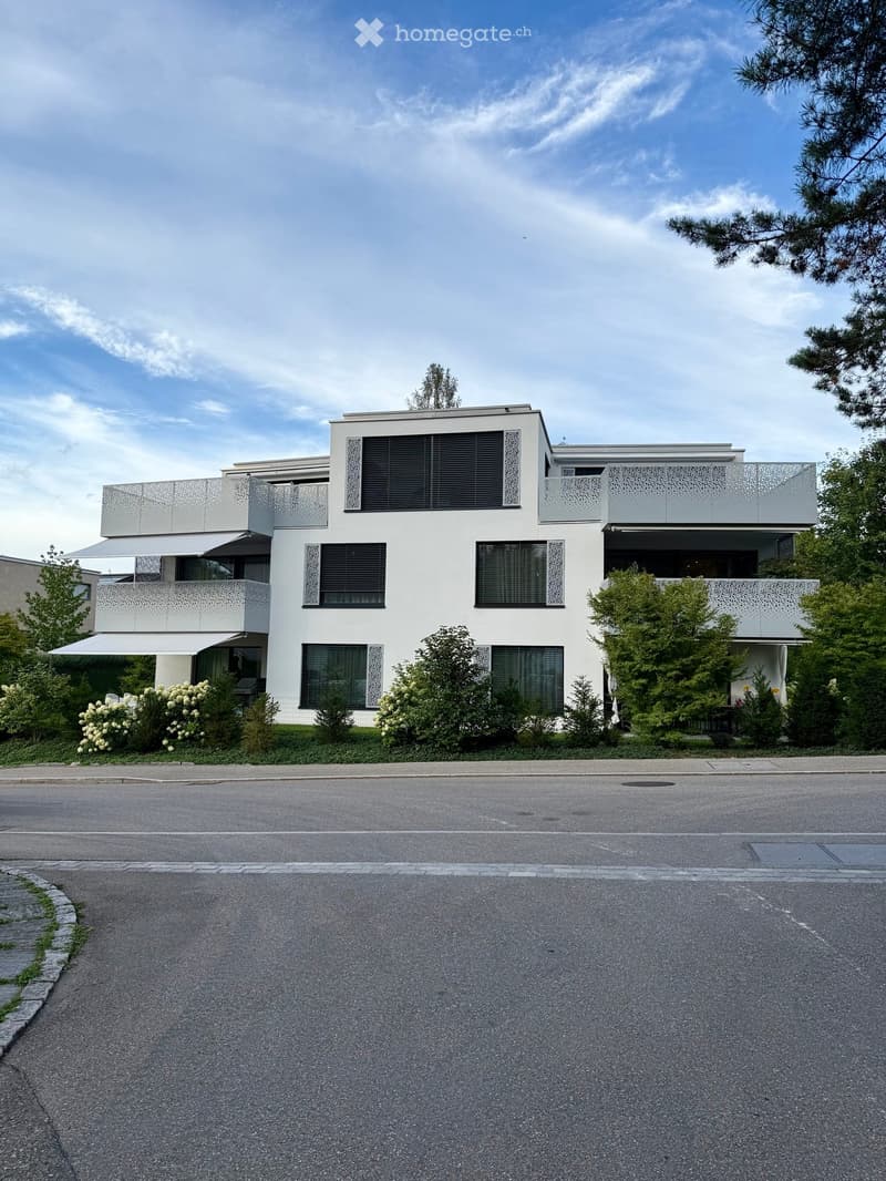 Modern two-story house with white exterior, large windows, balconies, and surrounding greenery.