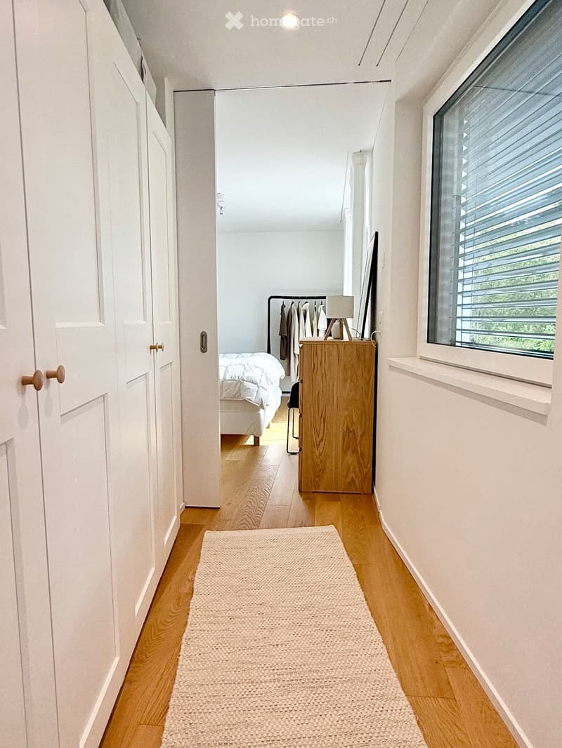 A narrow hallway with light wooden flooring, white walls, and a cream-colored runner rug. The hallway leads to a bedroom with a bed, nightstand, and clothing rack visible in the background.