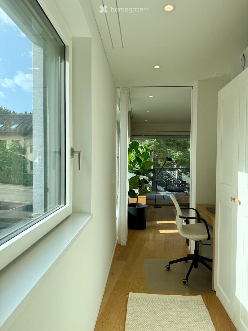 Bright, modern room with wooden flooring, large window, and a view of the garden. Features a desk with a laptop, a comfortable chair, a potted plant, and a floor lamp.
