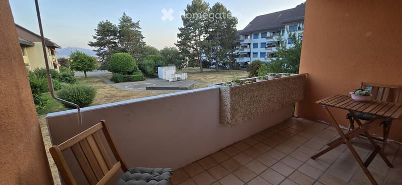 Balcony with tiled floor, wooden table, chairs, and a small bowl on the table. The view shows a landscaped area with trees and a building in the background.