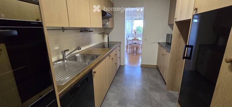 Modern kitchen with light wooden cabinets, stainless steel sink, oven, refrigerator, and dishwasher. Open to a dining area with a table and chairs.