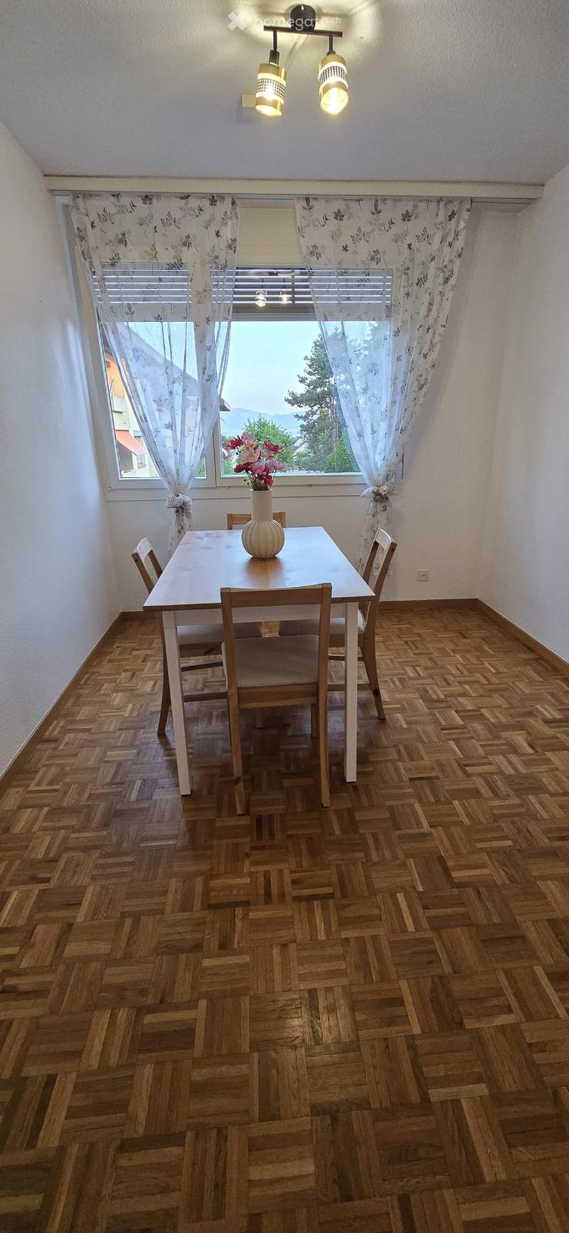 Dining room with wooden herringbone floor, white dining table, four chairs, and a vase with flowers on the table. Large window with floral curtains and a view of greenery.
