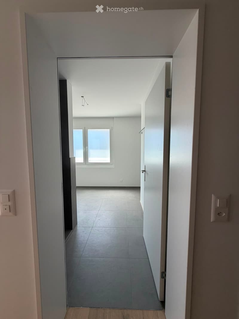 Empty hallway with tiled floor, white walls, and a door leading to a room with windows.