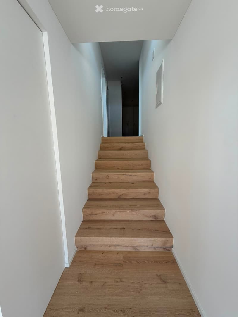 A modern, minimalist hallway with light wooden stairs leading upwards, white walls, and a polished wooden floor.