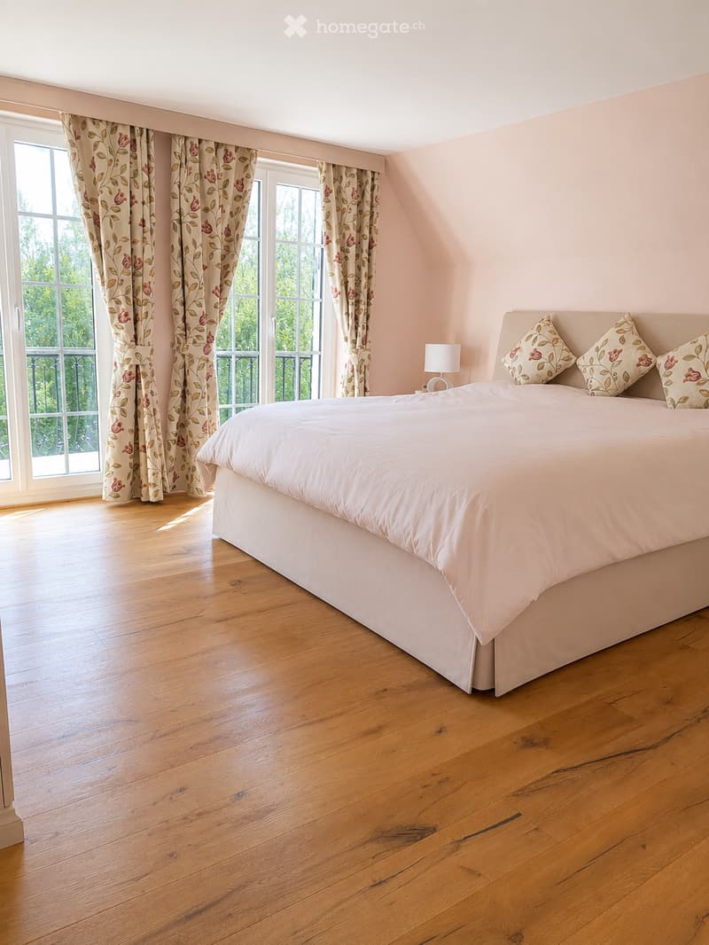 Spacious bedroom with wooden flooring, large windows with floral curtains, a neatly made bed with pillows, and a bedside lamp.