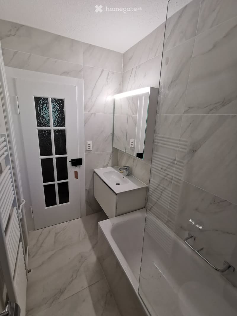 Modern bathroom with marble tiles, a white sink, bathtub, and a large mirror with built-in lighting. Features a frosted glass window and a white radiator.