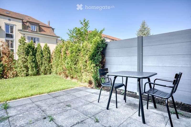 Garden area with a paved patio, a small black metal table, and two matching chairs, surrounded by greenery and a wooden fence.