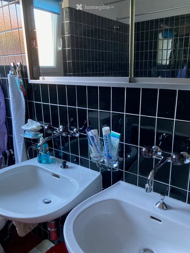 Modern bathroom with two white sinks, chrome faucets, toothbrush holders, toothpaste tubes, soap dispenser, and a towel on the side.