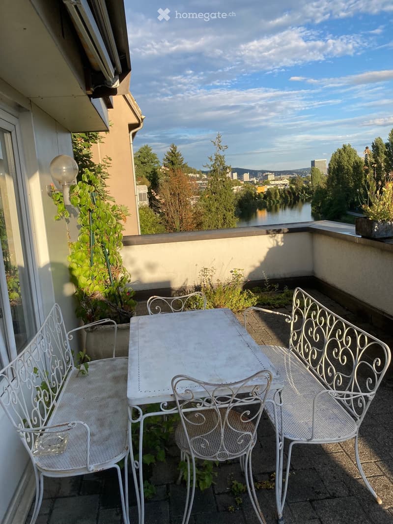 Balcony with a white metal table and chairs, overlooking a river and cityscape.
