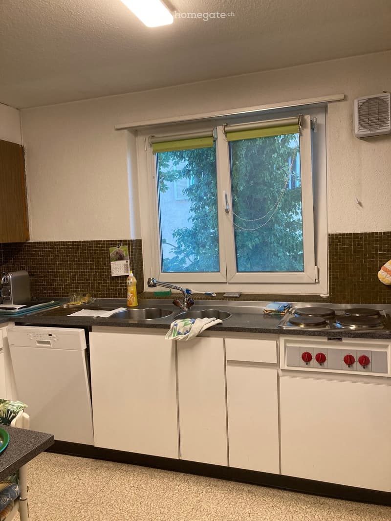 A compact kitchen with white cabinets, a black countertop, a sink with a faucet, a dishwasher, and a small electric stove. The room features tiled walls, a window with blinds, and various small items on the counter.