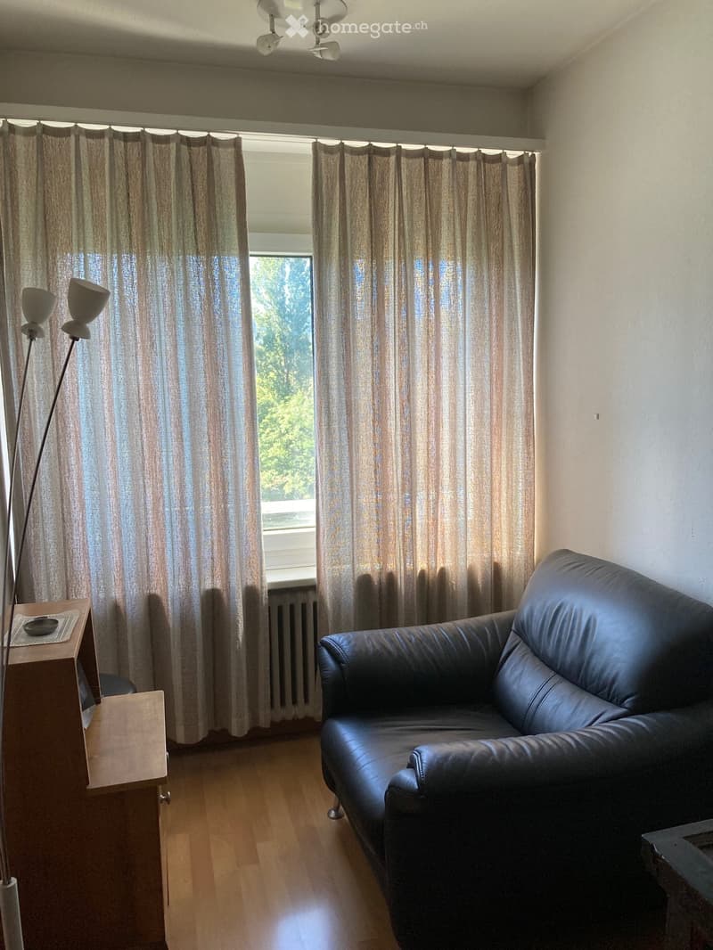 A modern living room with a black leather sofa, a wooden side table, and a tall floor lamp near the window with sheer curtains.