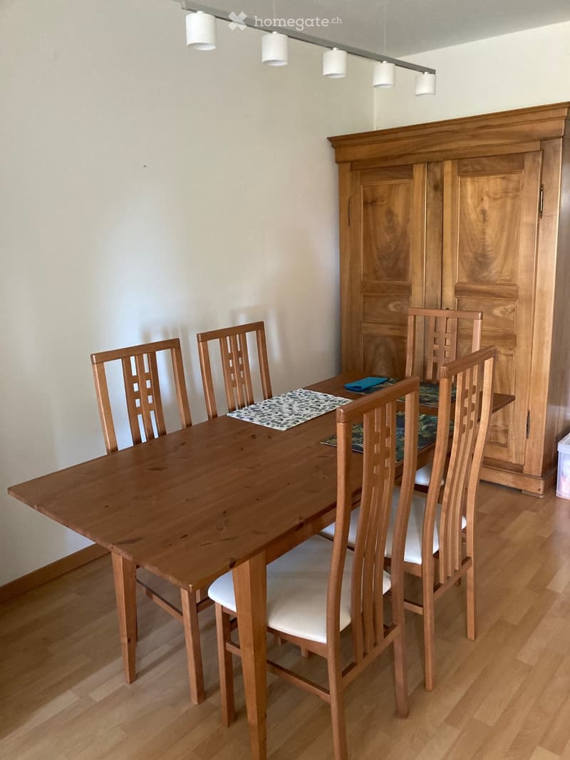 Dining room with a wooden dining table, four chairs with white cushions, and a wooden cabinet with glass doors.